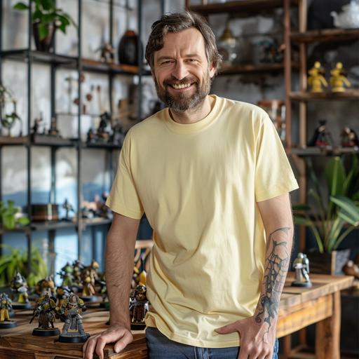 mockup of middle-aged man wearing a plain light yellow colored Bella-Canvas 3001 t-shirt, smiling, behind a table with dozens of dungeons and dragons miniatures