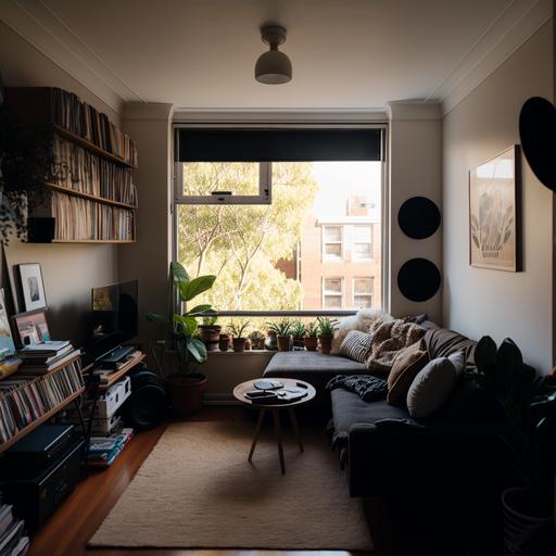 A living room of a small apartment, with a long horizontal window facing a busy street in Sydney. There is a black l-shaped couch below the window, and a wooden circle coffee table with black legs. Next to the couch is a cubed 4x4 wooden book shelf filled with records and books, with a record player and plant sitting on top of it. Between the book shelf and couch is a black lamp with a circle rattan lamp shade. The room is light filled, with dark grey carpet, is filled with plants and a cosy vibe.