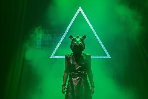A woman with a hippo head wearing a leather dress stands on a stage, with a triangular light coming from behind. Green smoke wafts through the air. A wirefram hologram forms the words 