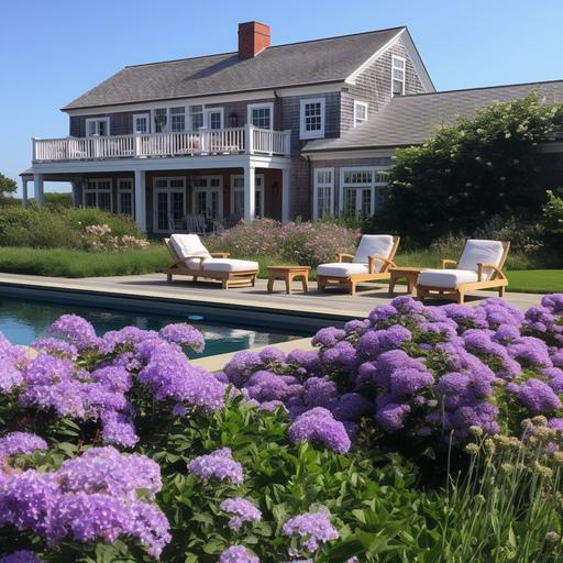 Gambrel style Nantucket house and separate pool house and large pool with summer patio furniture and a outdoor bbq. Ocean in the distance. Purple and white flowers in garden. In Tom Nevers Nantucket