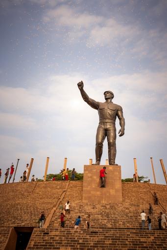 Make the statue a164 feet high and people climbing, clear sky in the background, picture taken from the sky, burkina Faso Falgs in the back --ar 2:3