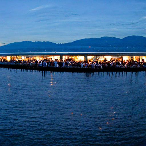 The longest chartuterie baord on White Rock, BC Canada's 1500 ft pier with 500 people dressed nicely drinking wine on the pier in cocktail style vibe. The charcuterie board is 500 ft long by 10 inches wide on a 500 foot table coered in black table cloths with walking room around and Jazz trio performing at the end of the pier and all enjoying the stunning backdrop of White Rock and Semiahmoo Bay photorealistic --v 3 --video