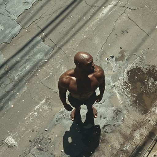 camera very high angle rainging bullet on the street a black man bald muscle is in the middle of the street standing