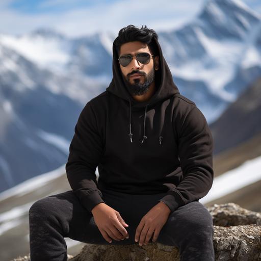 photoshoot of a 6 feet tall brown Indian man wearing high quality, plain black hoodie sitting on a rock. Background is in the snow capped mountains.