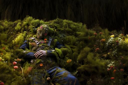 male torso lying on the ground covered with wildflowers and moss, one of his palms resting on his chest