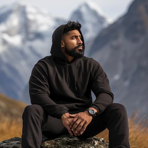 side profile photoshoot of a 6 feet tall brown Indian man wearing high quality, plain black hoodie sitting on a rock. Background is in the snow capped mountains.