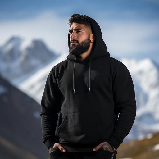 side profile photoshoot of a 6 feet tall rugged looking Indian man wearing high quality, plain black hoodie standing in style. Background is in the snow capped mountains.