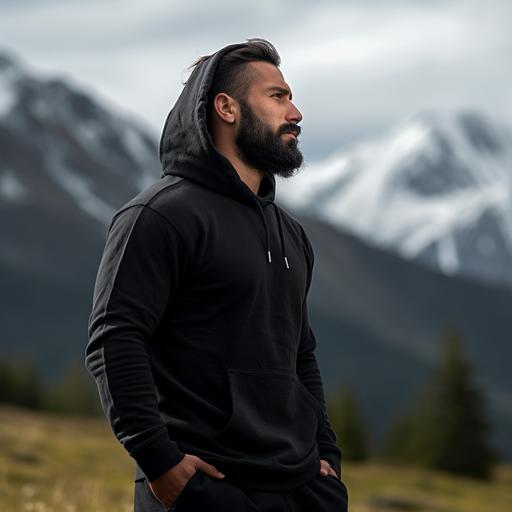 side profile photoshoot of a 6 feet tall rugged looking Indian man wearing high quality, plain black hoodie standing in the meadows with pine and snow capped mountain in the background.